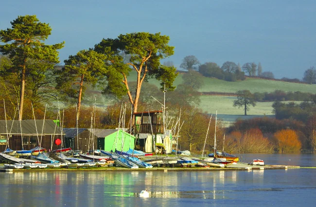 Small sailing dinghies drawn up on a bank at the edge of still water, with boat sheds and tall trees behind